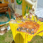Festivalstand mit gelbem Tischtuch, Federfächern, Treibholz und einer bemalten Holzplatte, im Hintergrund die Mother-Drum