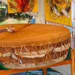 Die Mother-Drum aus Fell mit Schlägeln, auf dem Holzgestell im Zelt, dahinter gelbe Gemälde mit Federmotiven, davor ein Schaffell
