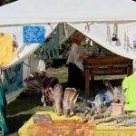 Weißes Festivalzelt mit gemalten Bildern und Federschmuck, darin spielt Gudrun die Mother-Drum, davor ein gelb gedeckter Tisch mit Federfächern