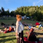 Gudrun steht mit Trommelschlägeln zwischen Teilnehmenden, die entspannt auf der Festivalwiese liegen, im Hintergrund Zelte und Wald